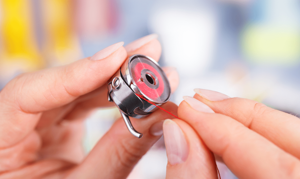 Close-up of a person’s hands threading a red bobbin into a metal bobbin case, preparing an embroidery machine for proper lower thread tension.